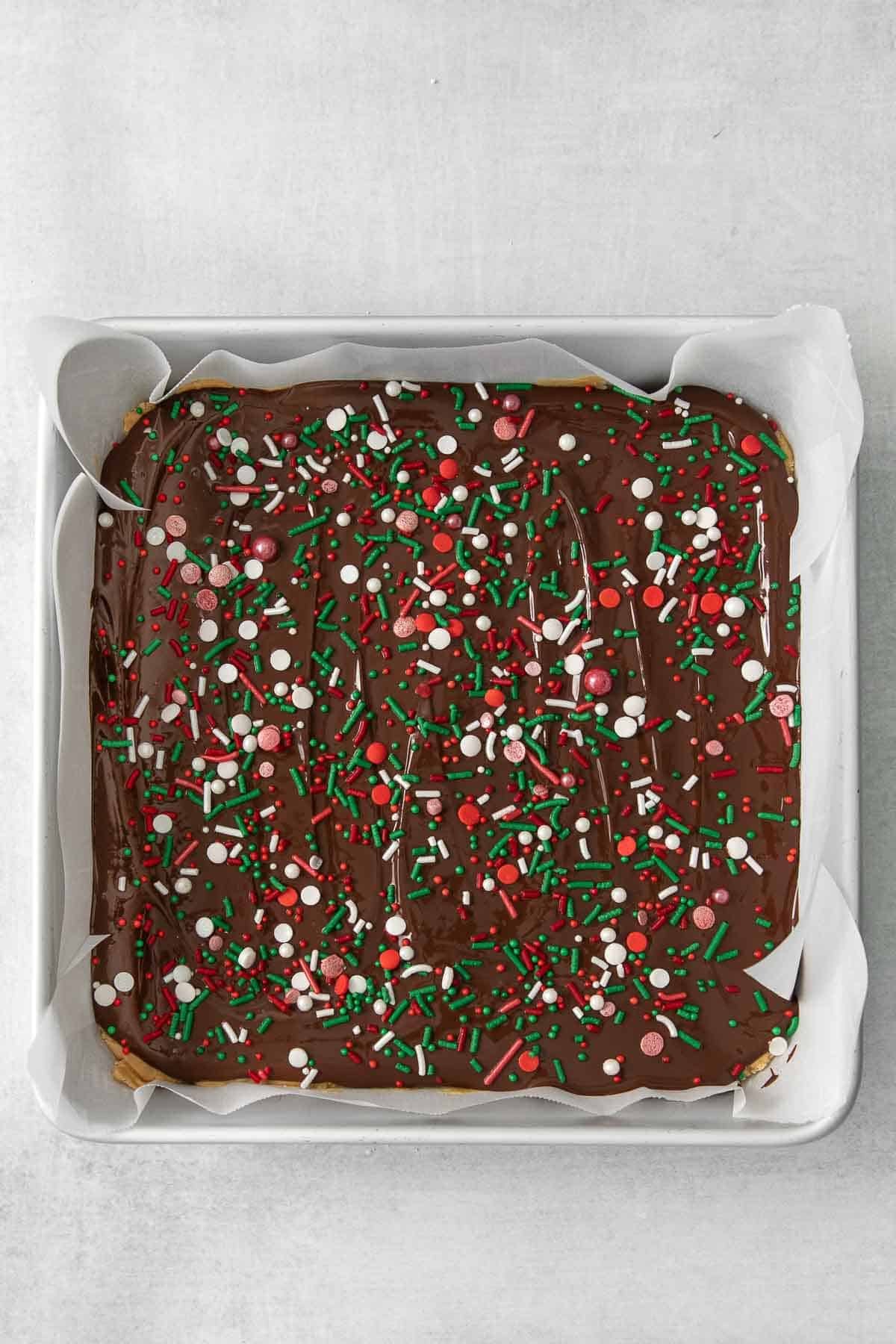 square metal baking dish with chocolate spread over a peanut butter mixture and topped with red, green and white sprinkles.