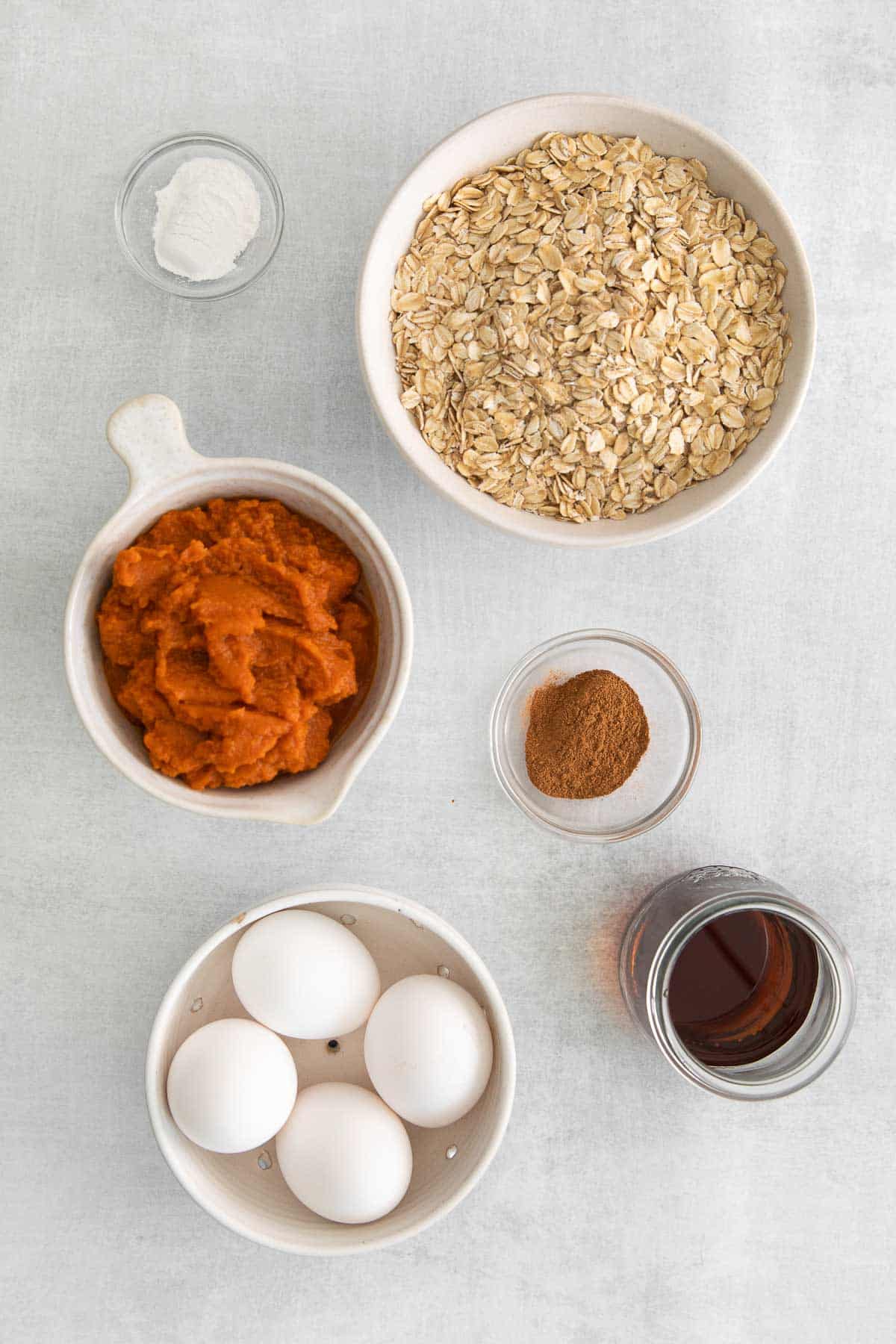 several white bowls with ingredients for pumpkin baked oatmeal - old fashioned oats, four eggs, pumpkin puree, cinnamon, and maple syrup.
