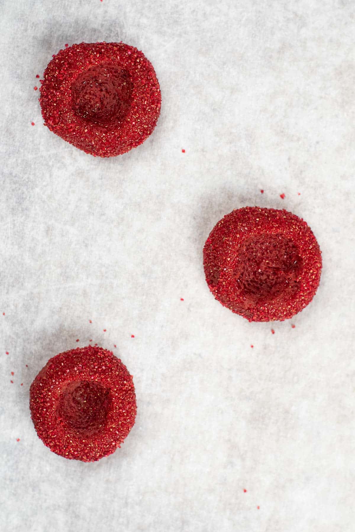 three sugar coated red velvet cookie dough with a thumbprint hole in each center.