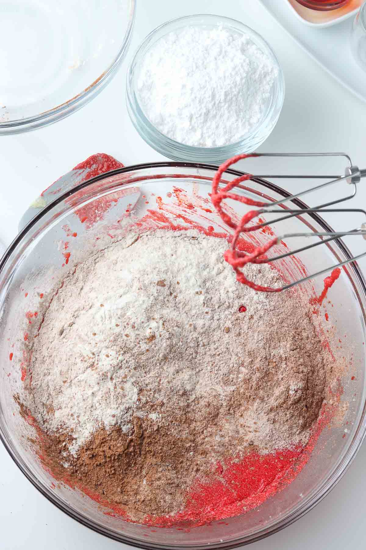 glass mixing bowl with red cookie batter with cocoa powder and flour on top.