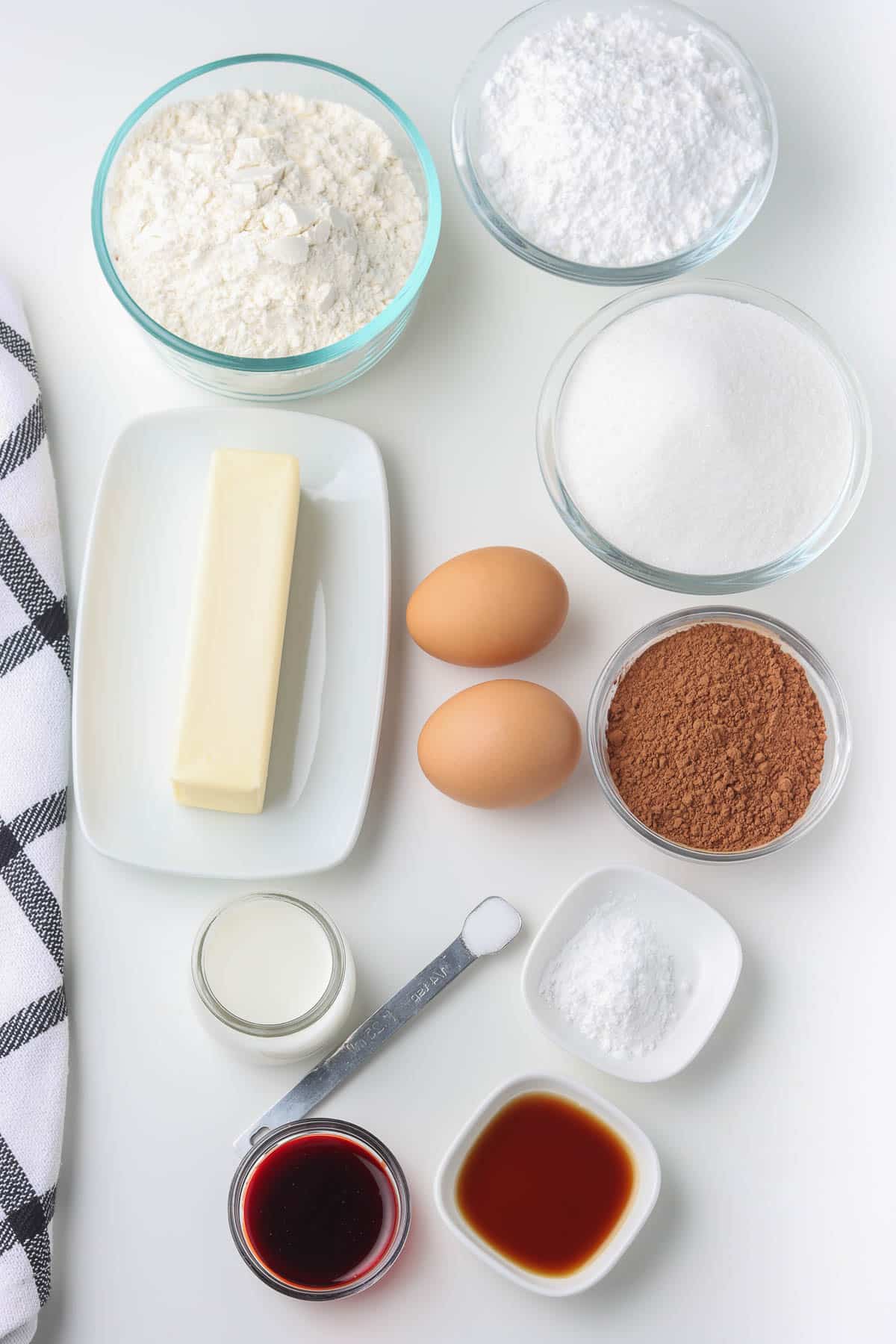 several white bowls with ingredients for red velvet cookies - flour, sugar, cocoa, vanilla extract, stick of butter, baking powder, milk, two eggs, and powdered sugar.