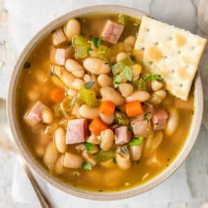 closeup of a bowl full of ham and bean soup with diced carrots and celery and a saltine cracker on the side of the bowl.