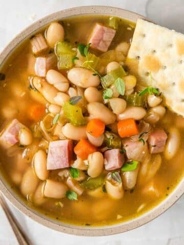 closeup of a bowl full of ham and bean soup with diced carrots and celery and a saltine cracker on the side of the bowl.