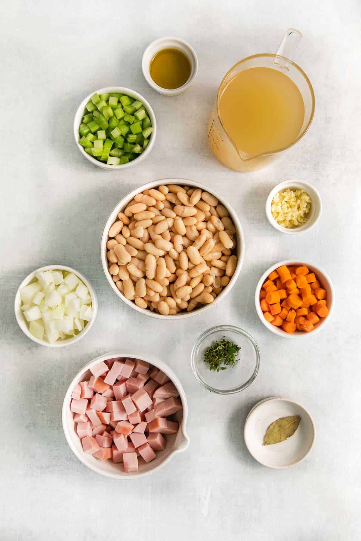 several small bowls with ingredients for white bean and ham soup - Cannellini beans, diced ham, onion, celery, carrots, chicken broth, garlic, oilve oil and bay leaf.