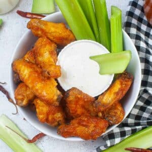 several buffalo chicken wings in a white bowl with celery sticks and a small bowl of ranch dressing.