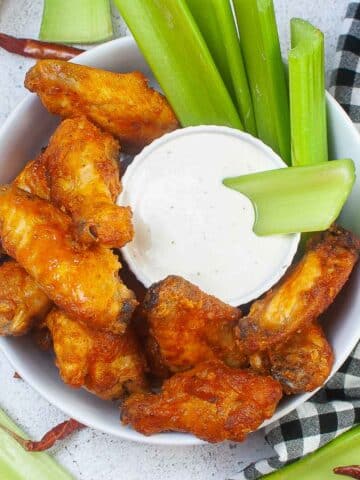 several buffalo chicken wings in a white bowl with celery sticks and a small bowl of ranch dressing.