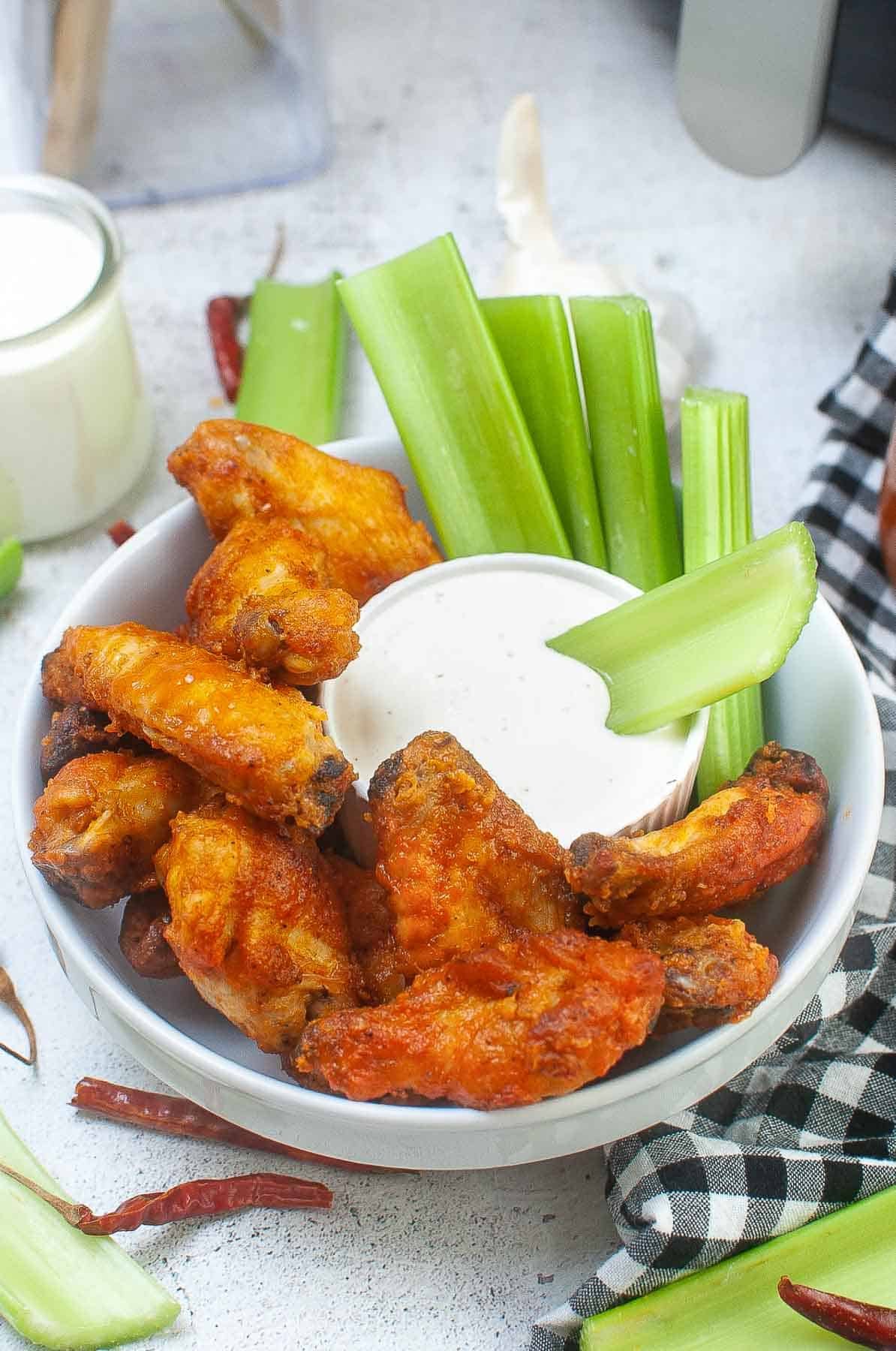 closeup of several buffalo chicken wings in a white bowl with celery sticks and a small bowl of ranch dressing.