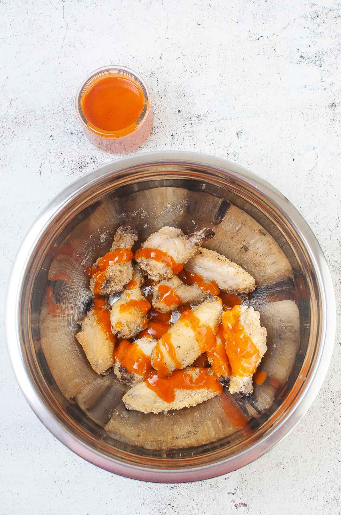 silver mixing bowl with chicken wings being tossed with buffalo sauce.