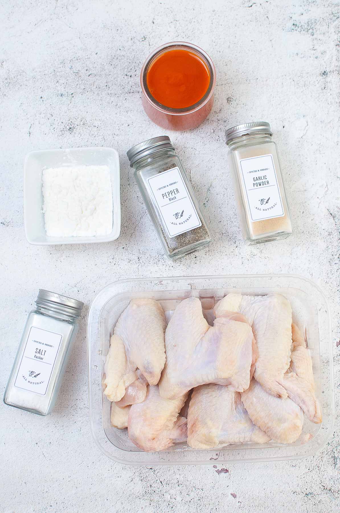 ingredients for buffalo wings - container of uncooked chicken wings, salt, pepper, garlic powder, buffalo sauce and corn starch.