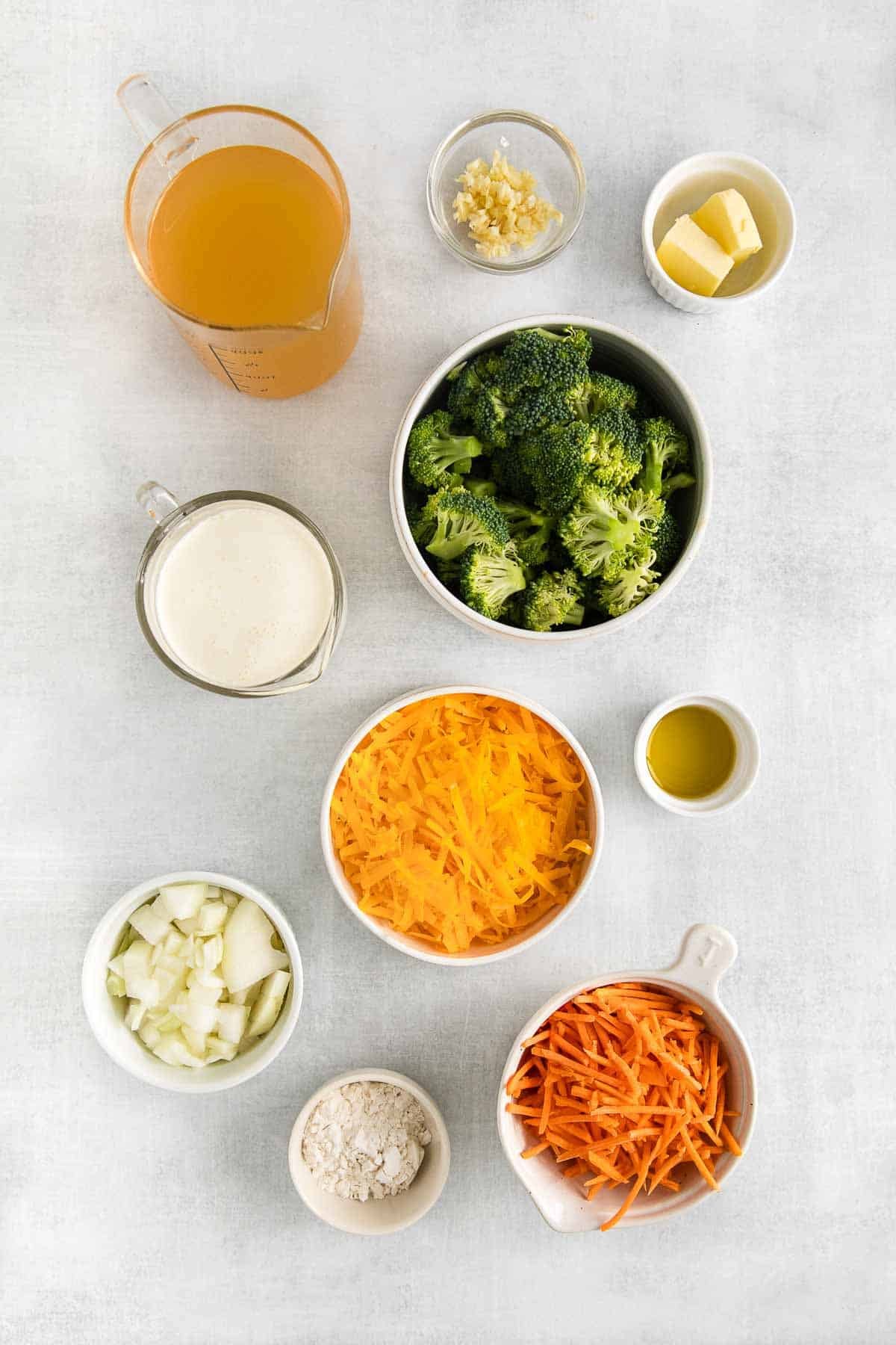 several small bowls with ingredients for broccoli cheddar cheese soup - broccoli florets, shredded cheddar cheese, shredded carrots, diced onions, chicken broth, cream, butter and four.