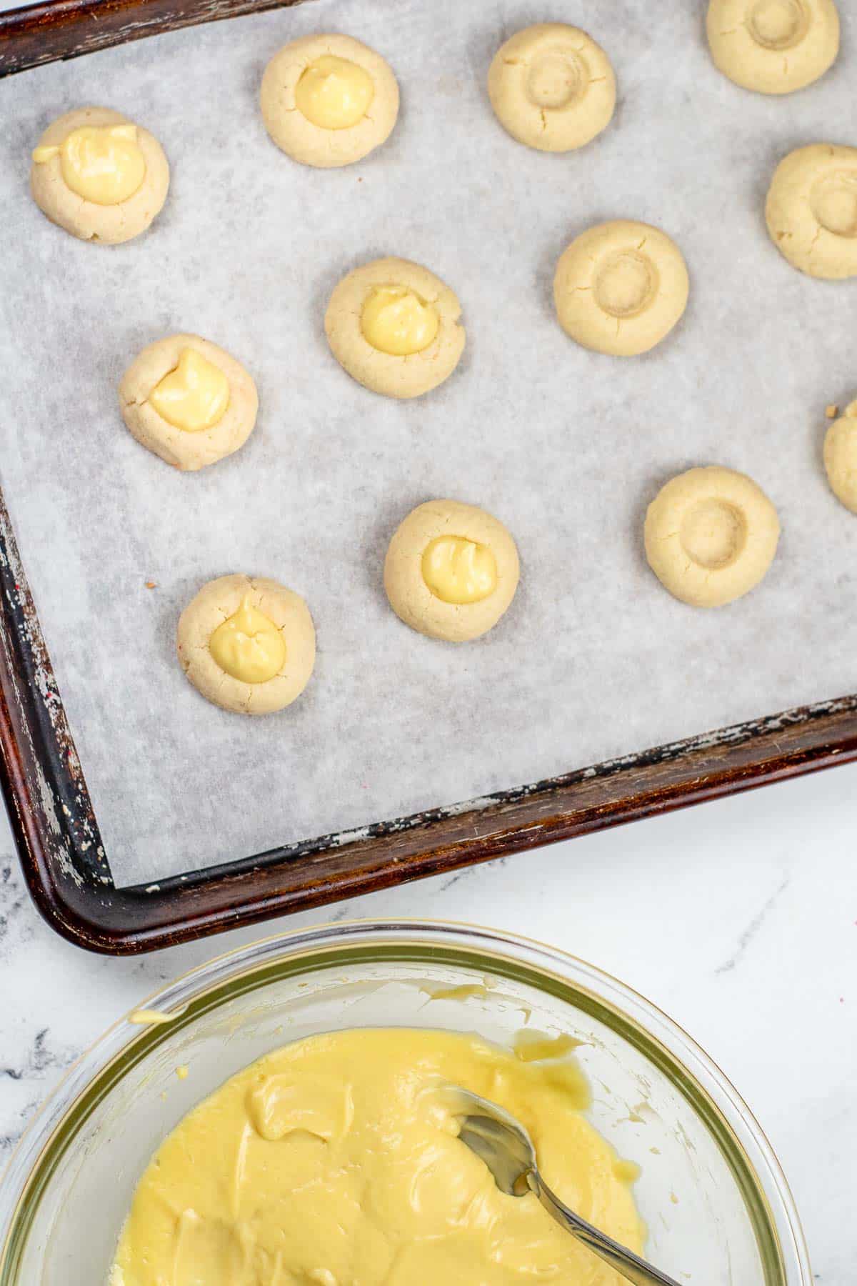 a baking sheet with several shortbread thumbprint cookies being filled with cheesecake filling in the center.