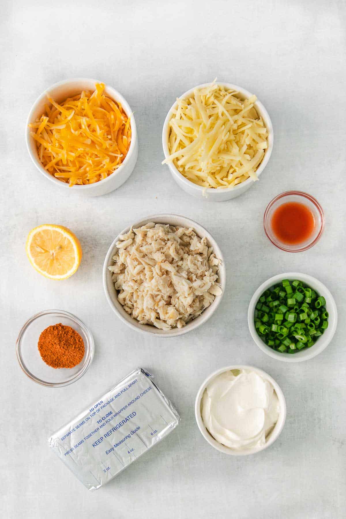several small bowls with ingredients for crab dip - lump crab meat, cream cheese, sour cream, shredded cheeses, old bay seasoning, and half a lemon.