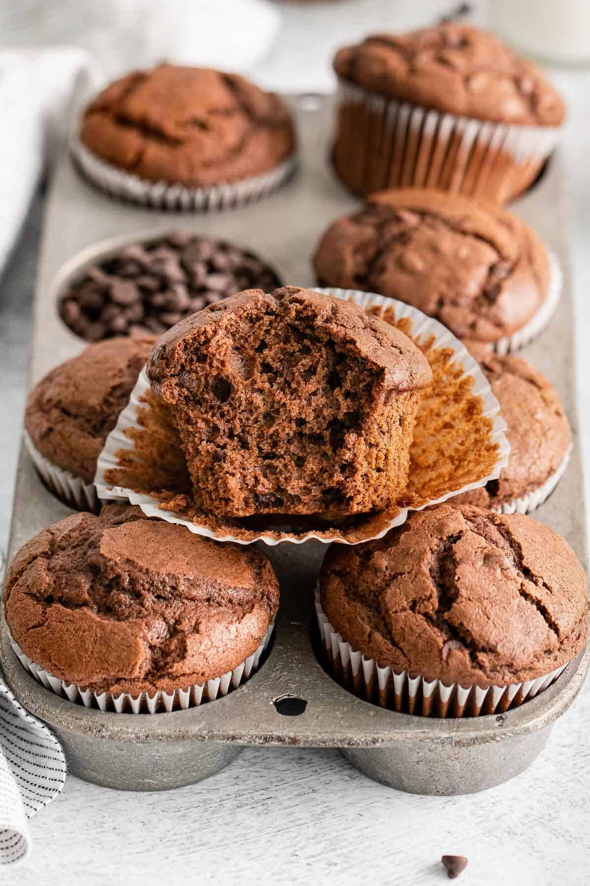 chocolate muffins in a muffin tin with one on top opened and a bite taken out.