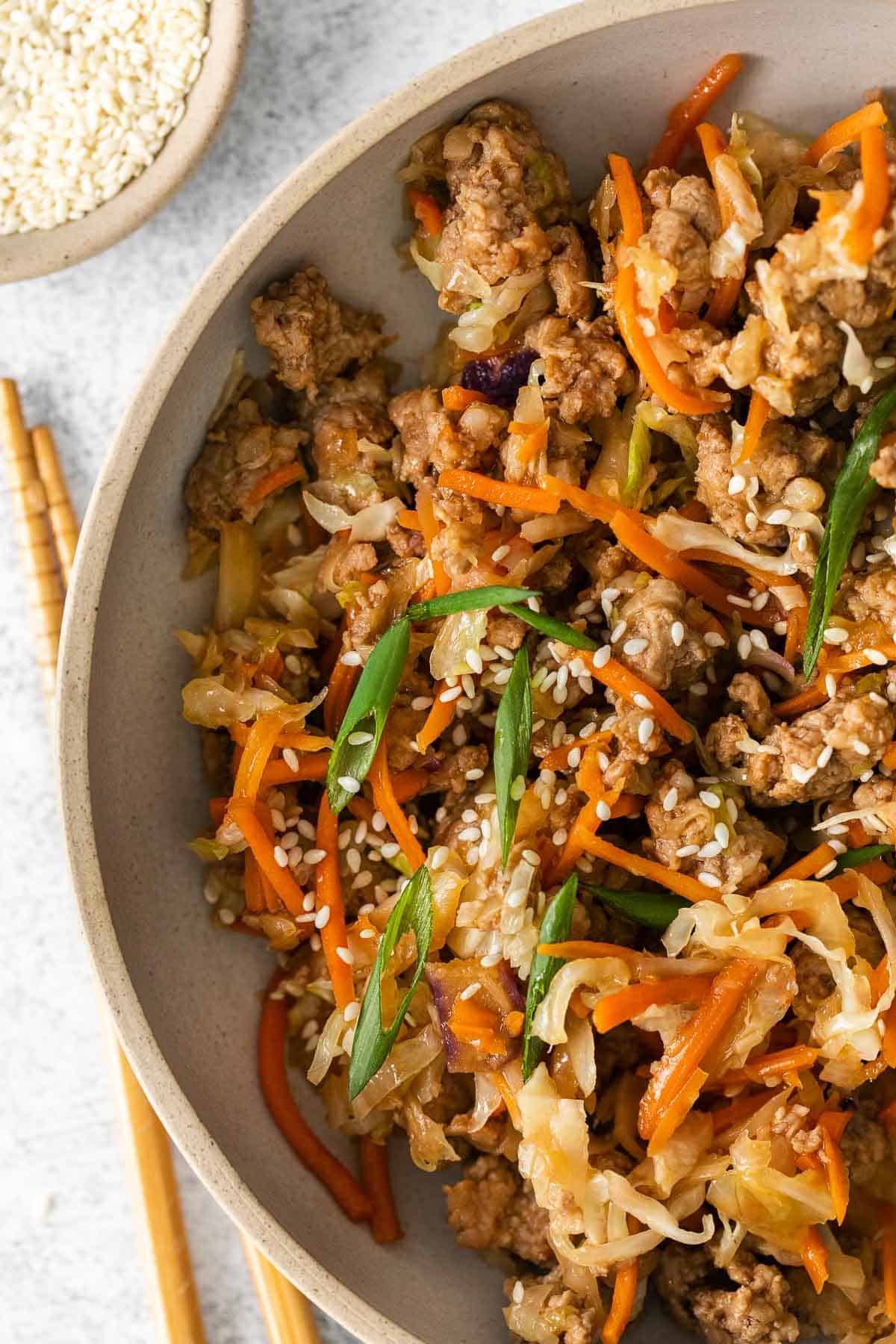 closeup of egg roll in a bowl with ground pork, shredded cabbage and carrots topped with sesame seeds and sliced scallions.