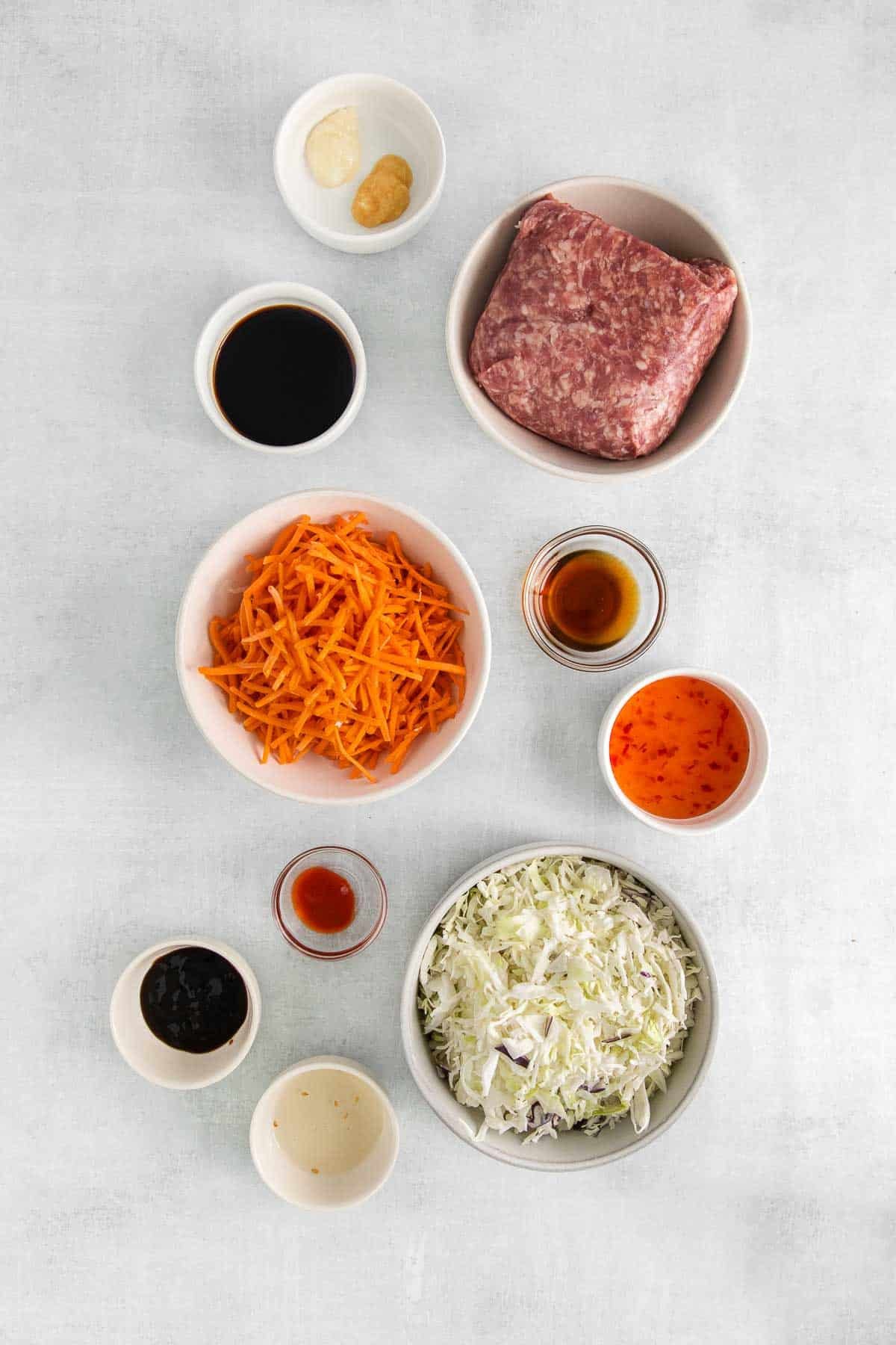 several white bowls with ingredients for egg roll in a bowl - ground pork, shredded cabbage, shredded carrots, soy sauce, sweet chili sauce, hoisin sauce, sesame oil, garlic and ginger.