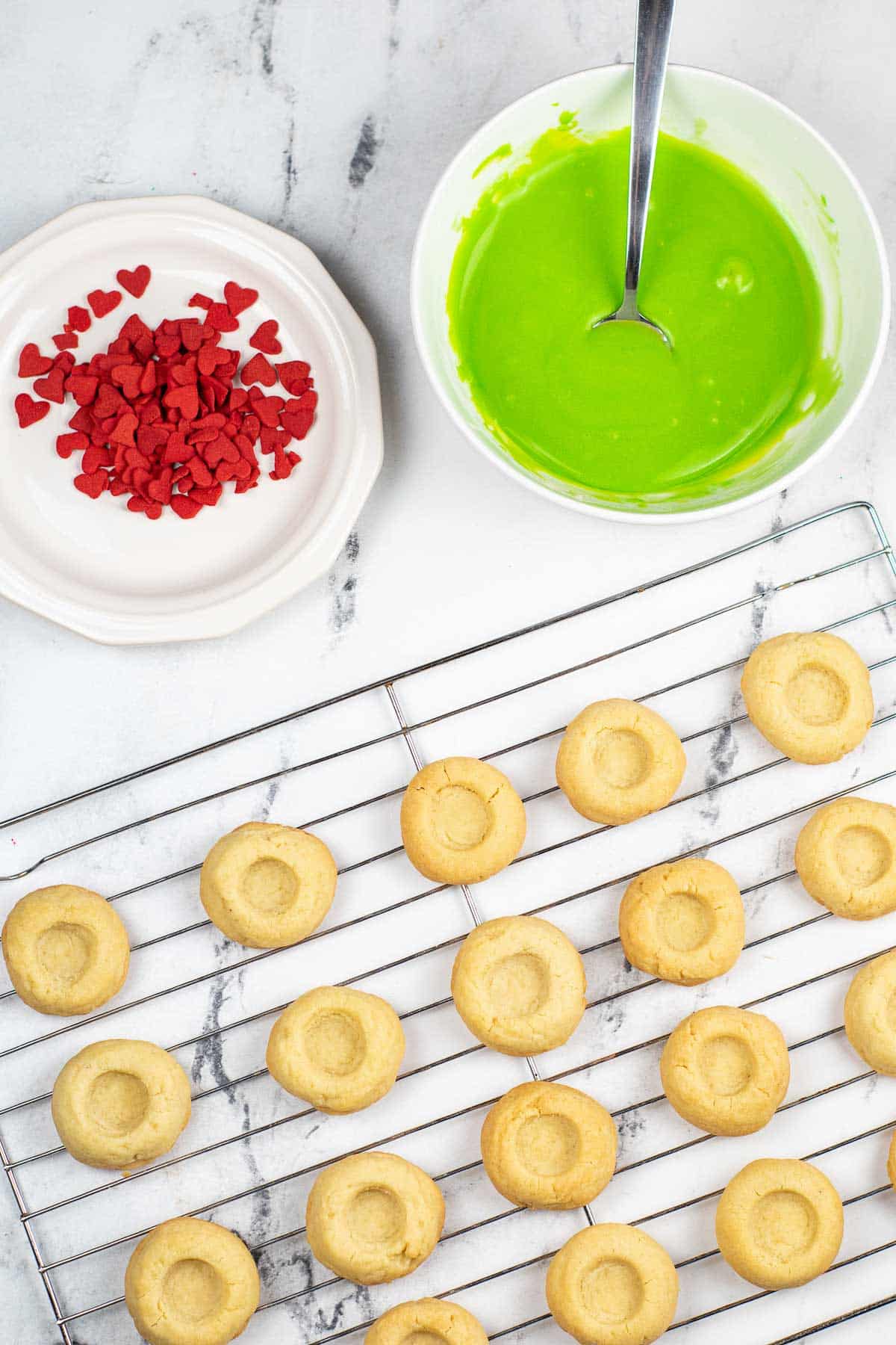 several buttery thumbprint cookies on a wire cooling rack with a bowl of green icing and little red candy hearts.