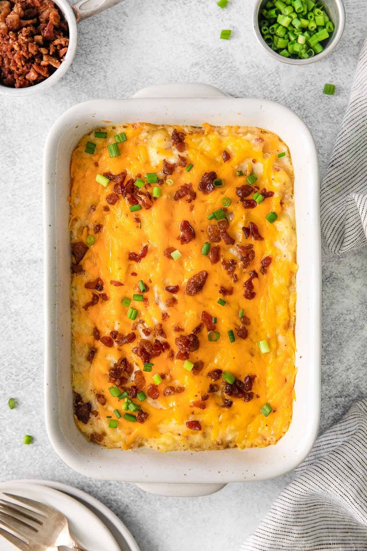 white baking dish with twice baked potato casserole topped with cheese, bacon bits and sliced scallions.