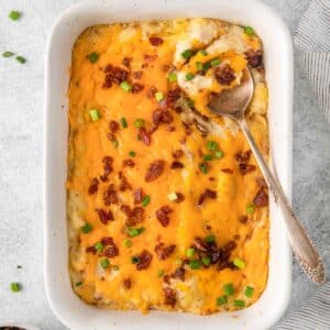rectangle baking dish with cheesy potato casserole with sliced scallions and bacon and a serving spoon on the side of the dish.