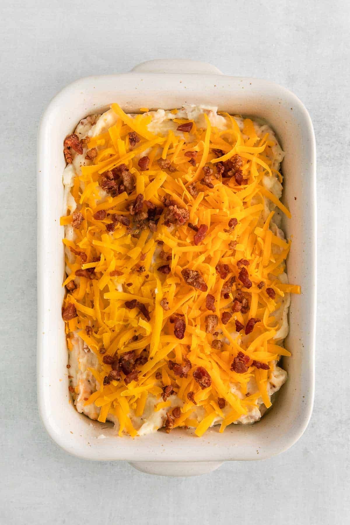 white rectangle baking with mashed potatoes topped with shredded cheese and bacon crumbles.
