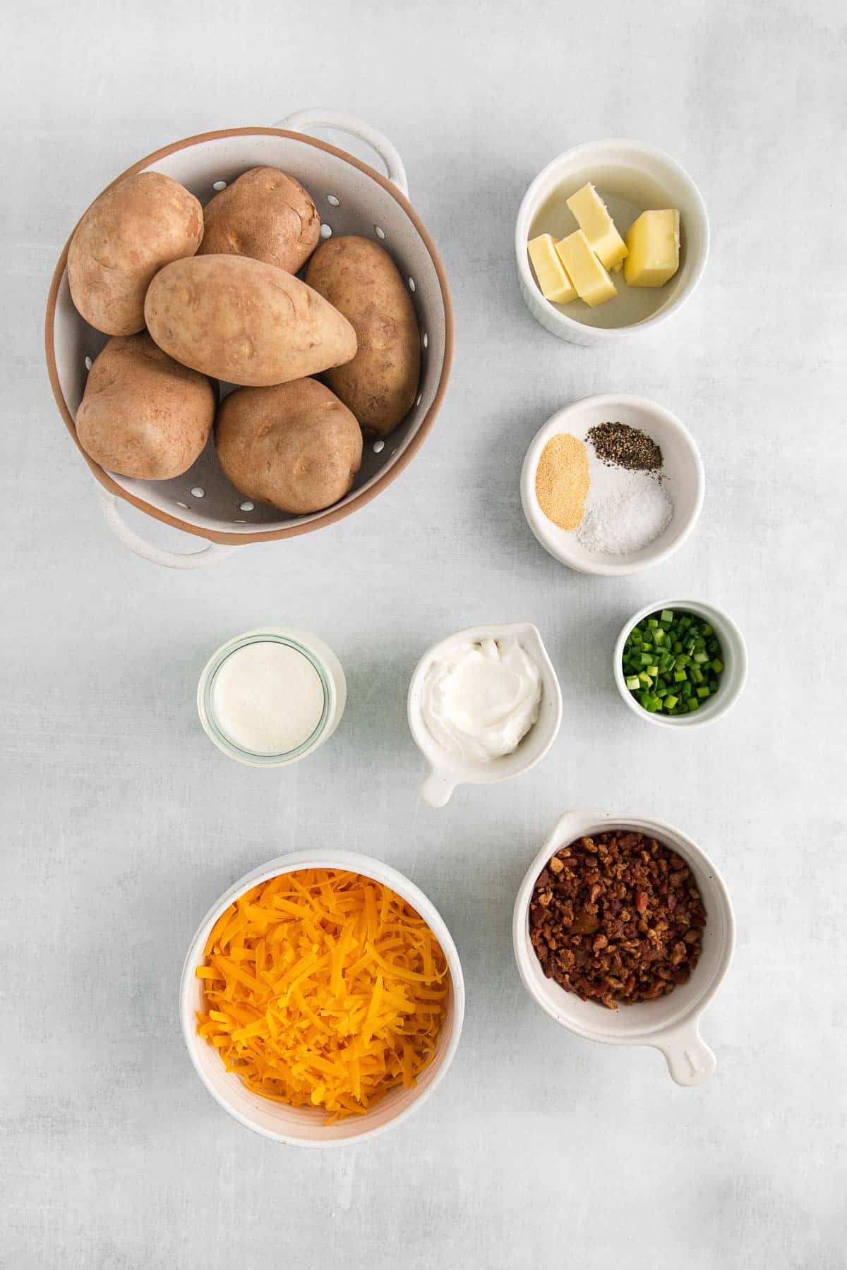 several bowls of ingredients for loaded potato casserole - russet potatoes, shredded cheese, bacon bits, butter, sour cream, scallions and spices.