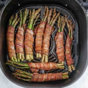 nine bundles of 3 or 4 asparagus spears wrapped in bacon in an air fryer basket.