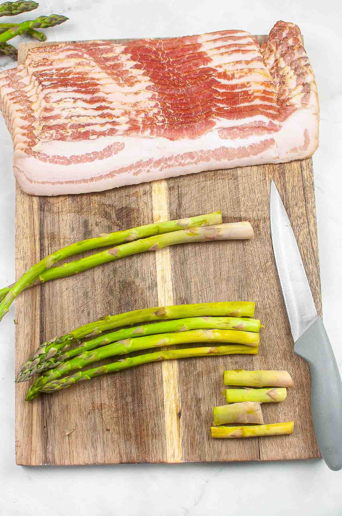 raw bacon slices and four spears of asparagus on a wood cutting board with the ends cut off.