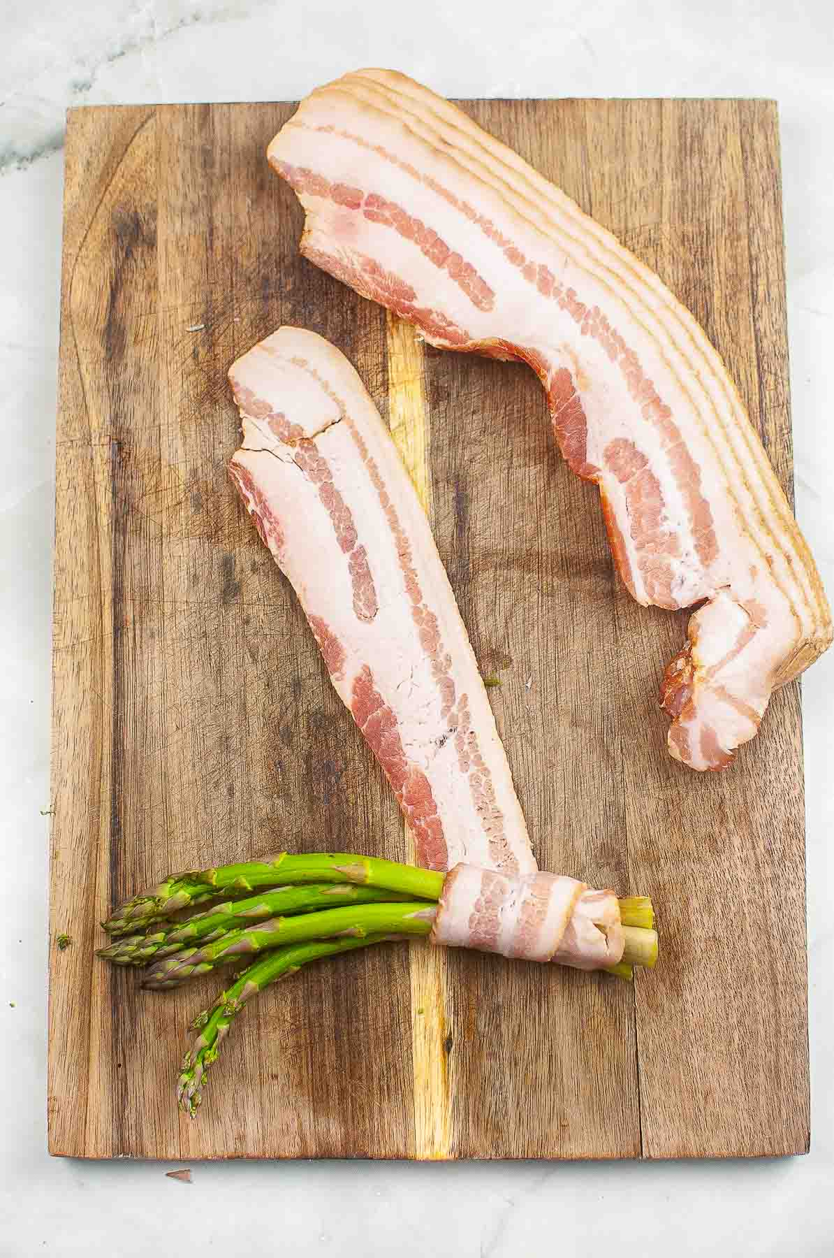 wood cutting board with four spears of asparagus being wrapped with raw bacon.