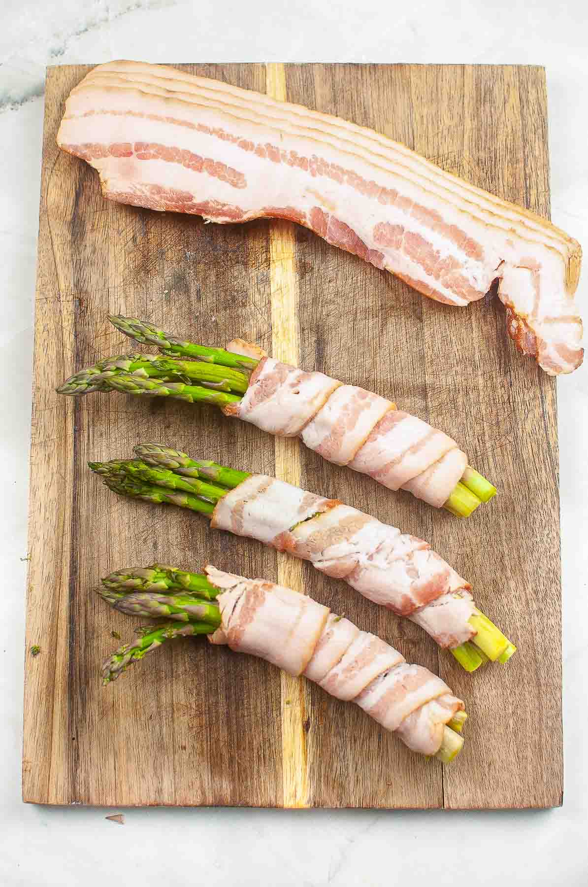 wood cutting board with three bundles of four asparagus spears wrapped in raw bacon.