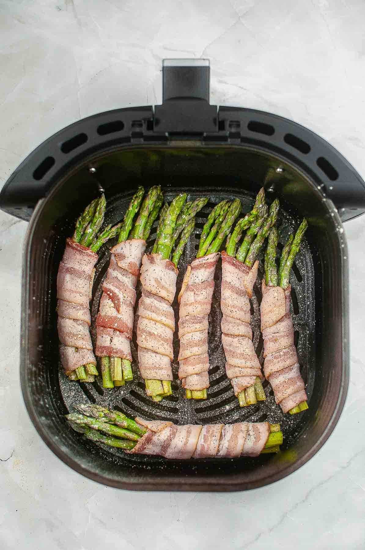 black air fryer basket with seven asparagus bundles wrapped in raw bacon.