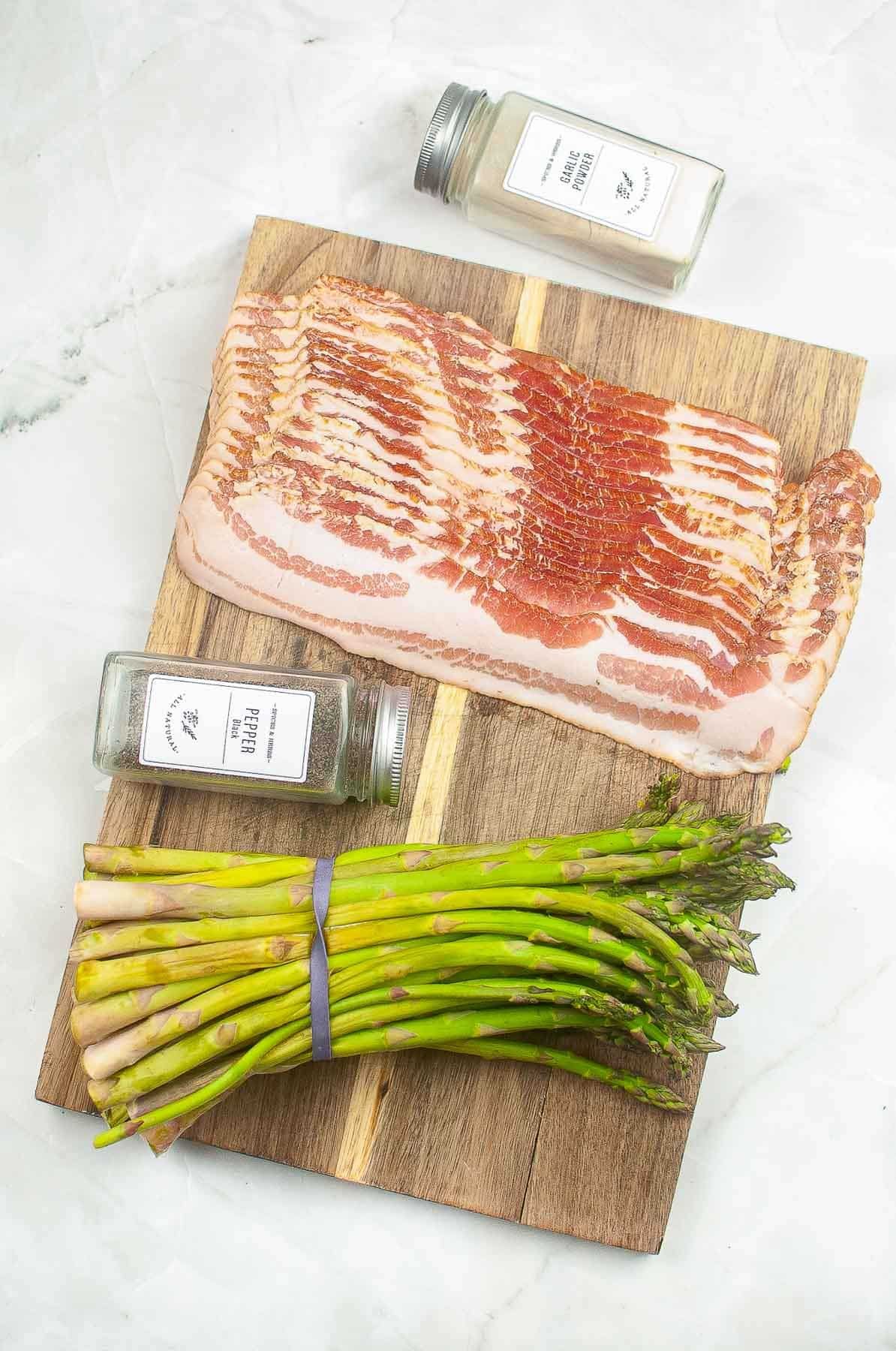 wood cutting board with one bunch of asparagus, a pound of sliced bacon and spice bottles of garlic powder and black pepper.