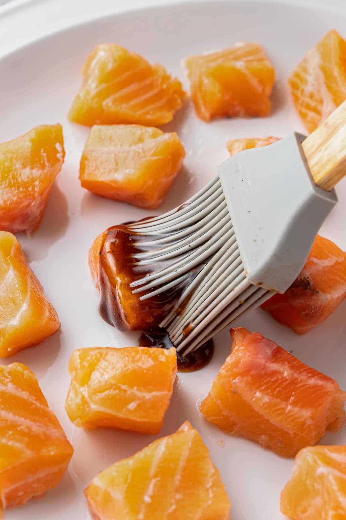 raw salmon cubes with a soy sauce mixture being brushed on with a gray brush.