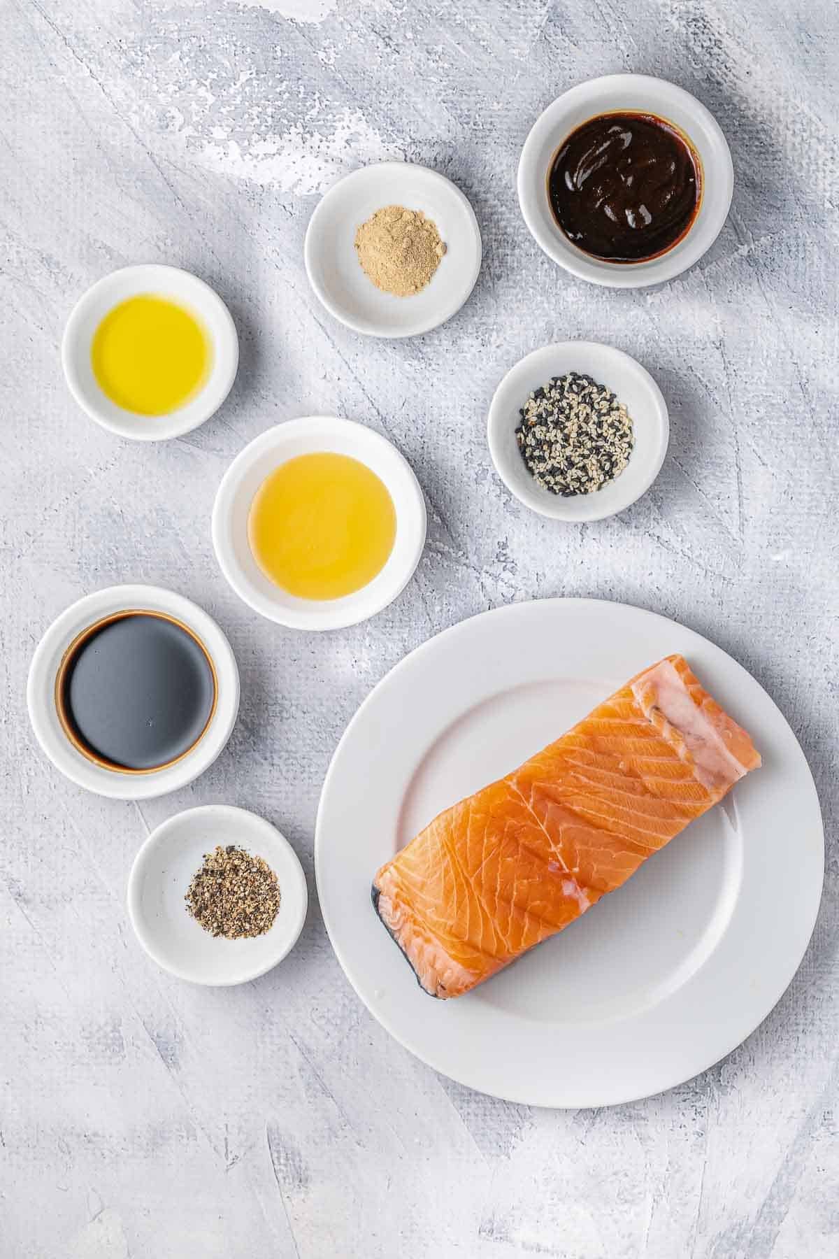 several small white bowls with ingredients for salmon bites - raw salmon, soy sauce, teriyaki sauce, olive oil, honey and garlic powder.