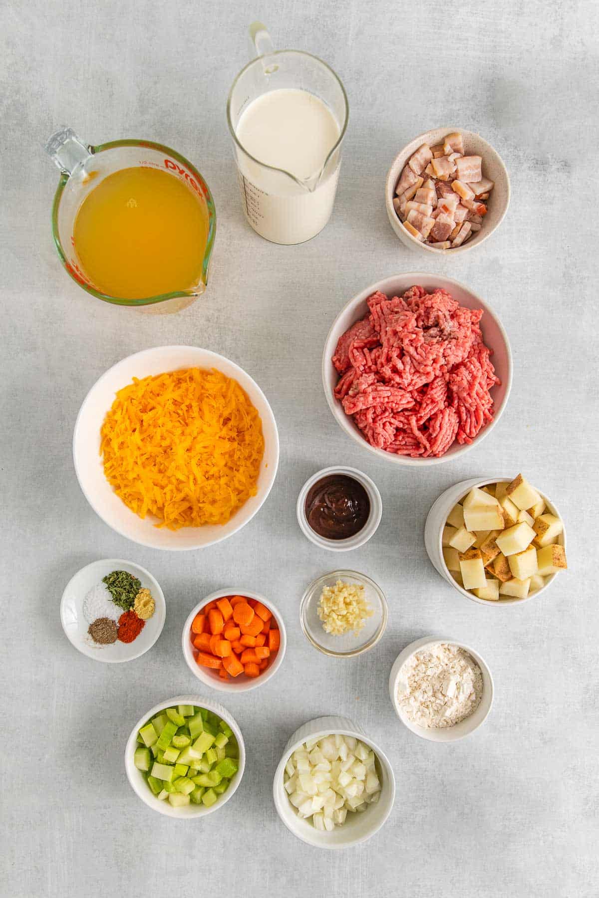 several small bowls with ingredients for cheeseburger soup - raw ground beef, bacon, diced potatoes, carrots, celery and onions, flour, cream, broth and shredded cheese.