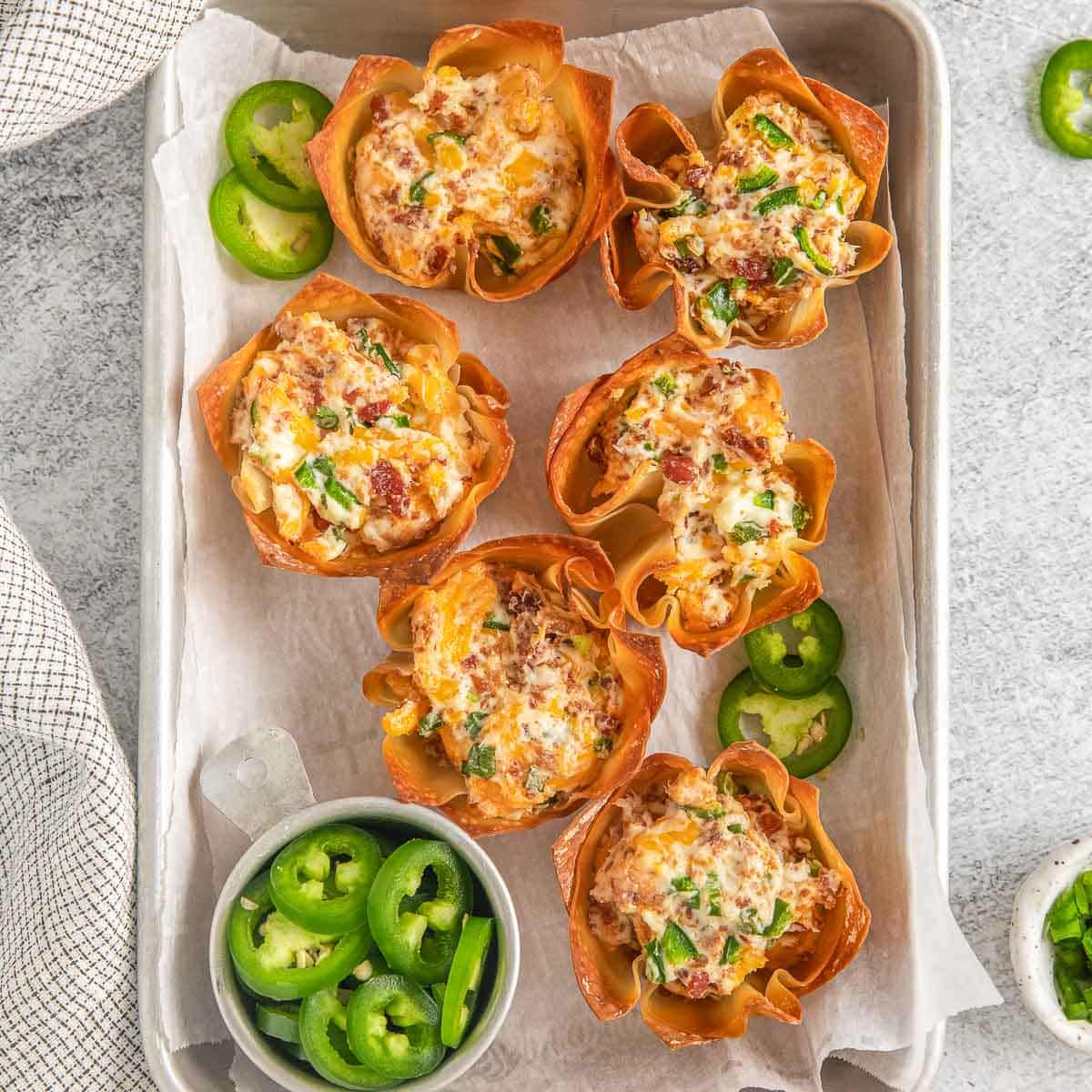 six jalapeno popper wonton cups on a small baking sheet lined white parchment paper.