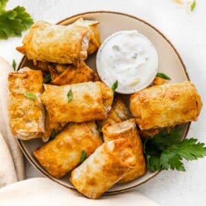 White plate of air fryer egg rolls topped with cilantro and a bowl of ranch on the side.