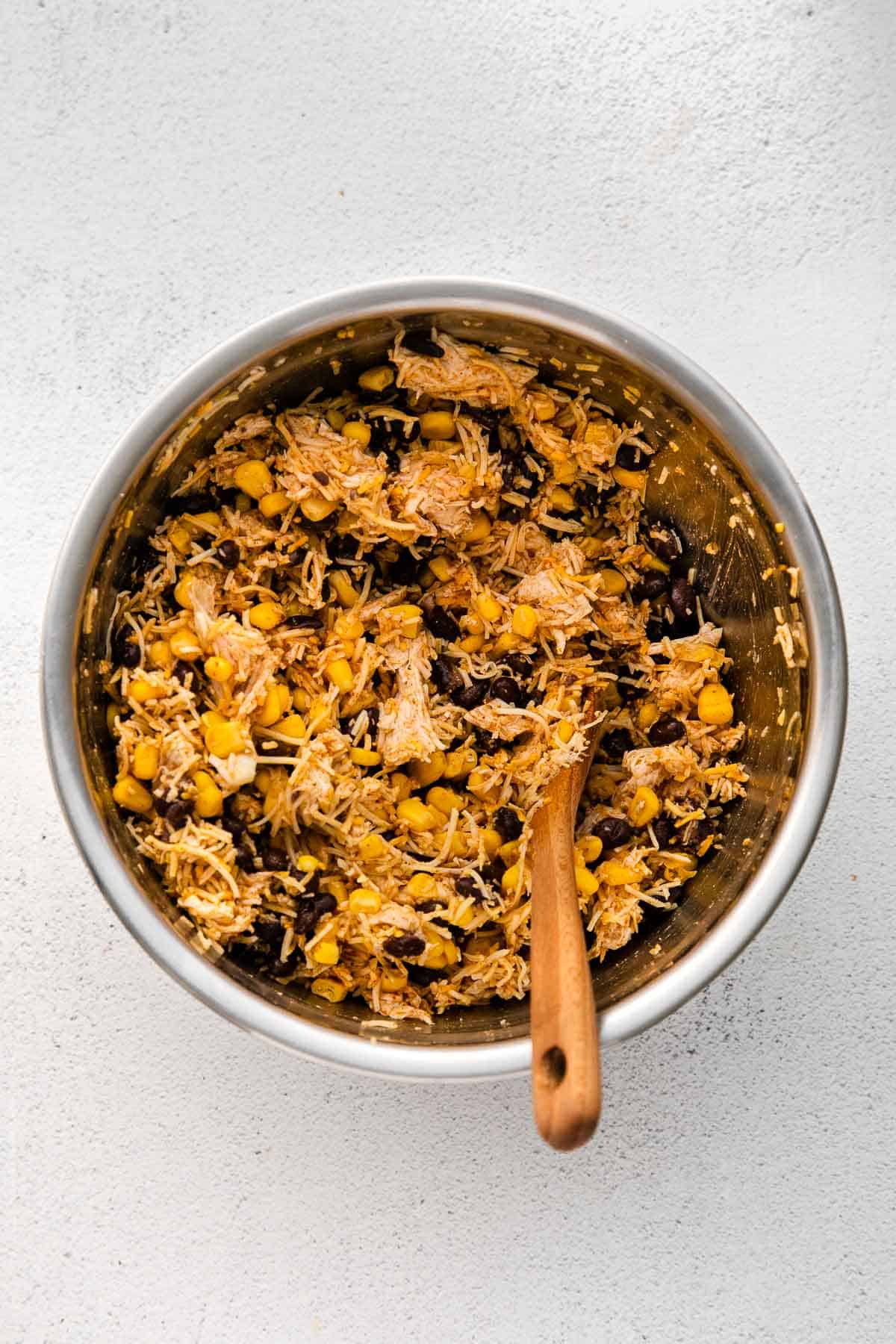 Medium mixing bowl of egg roll filling ingredients mixed together - chicken, cheese, beans, corn, chili powder, garlic powder, cumin, and paprika.