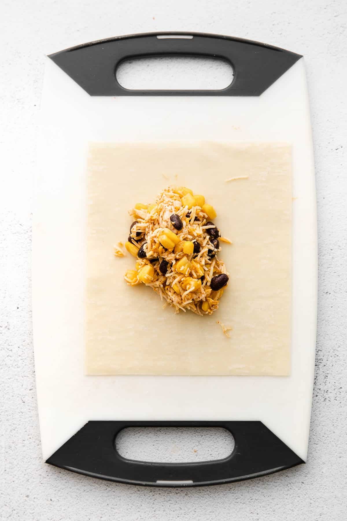 Cutting board with open egg roll wrapper with chicken, black beans, corn and shredded cheese mixture on top.