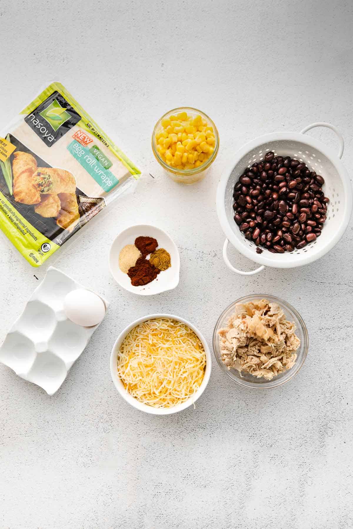 Several small bowls with ingredients for Air Fryer Southwestern Egg Rolls - chicken, black beans, corn, shredded cheese, egg, egg roll wrappers and seasonings.