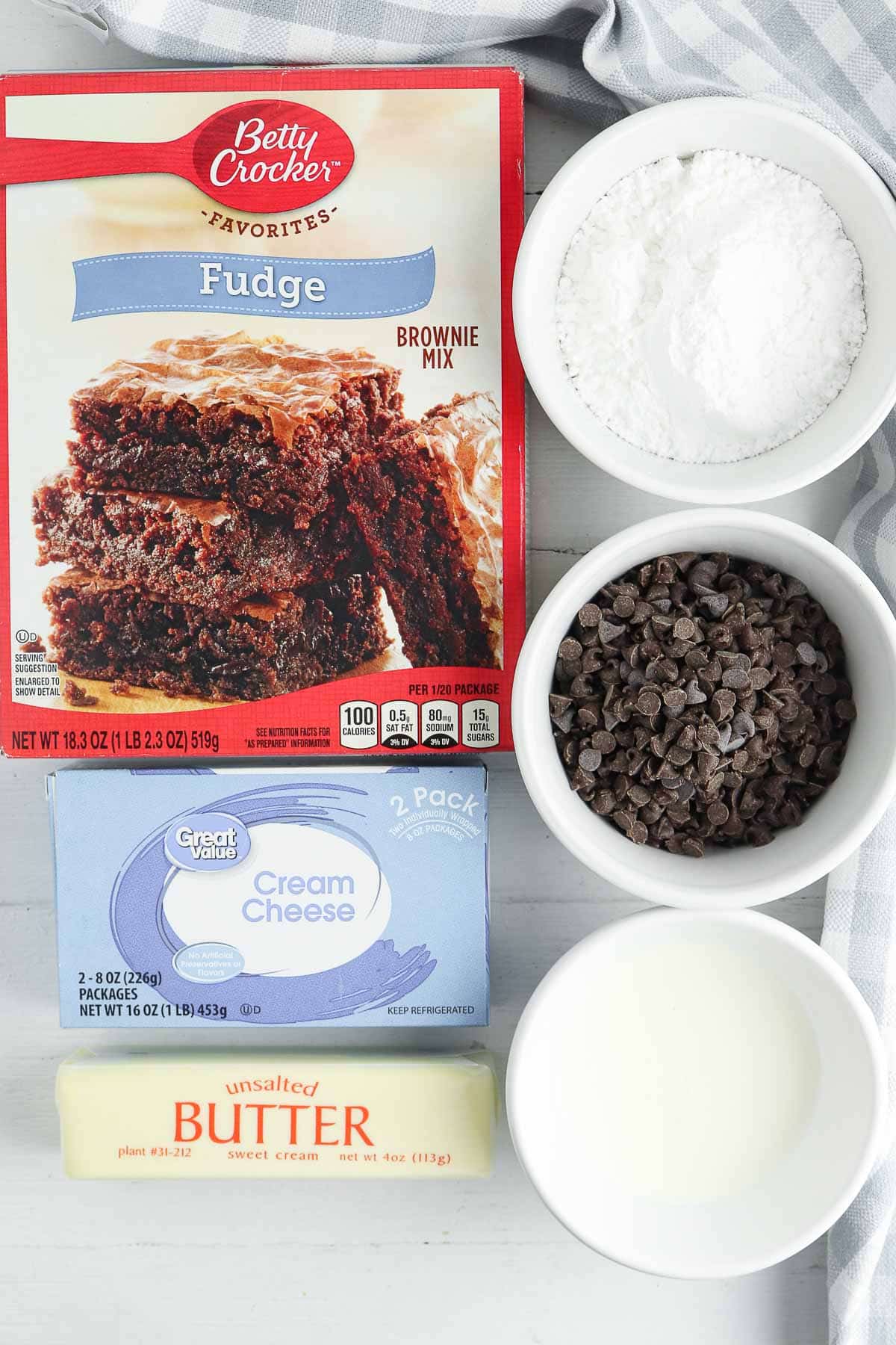brownie mix, stick of butter, block of cream cheese and white bowls with milk, powdered sugar and mini chocolate chips.