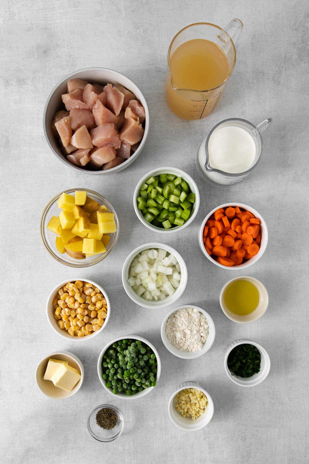 several small bowls with ingredients for chicken pot pie soup - raw diced chicken, chicken broth, milk, corn, onion, peas, carrots, butter, garlic, flour, oil and thyme.