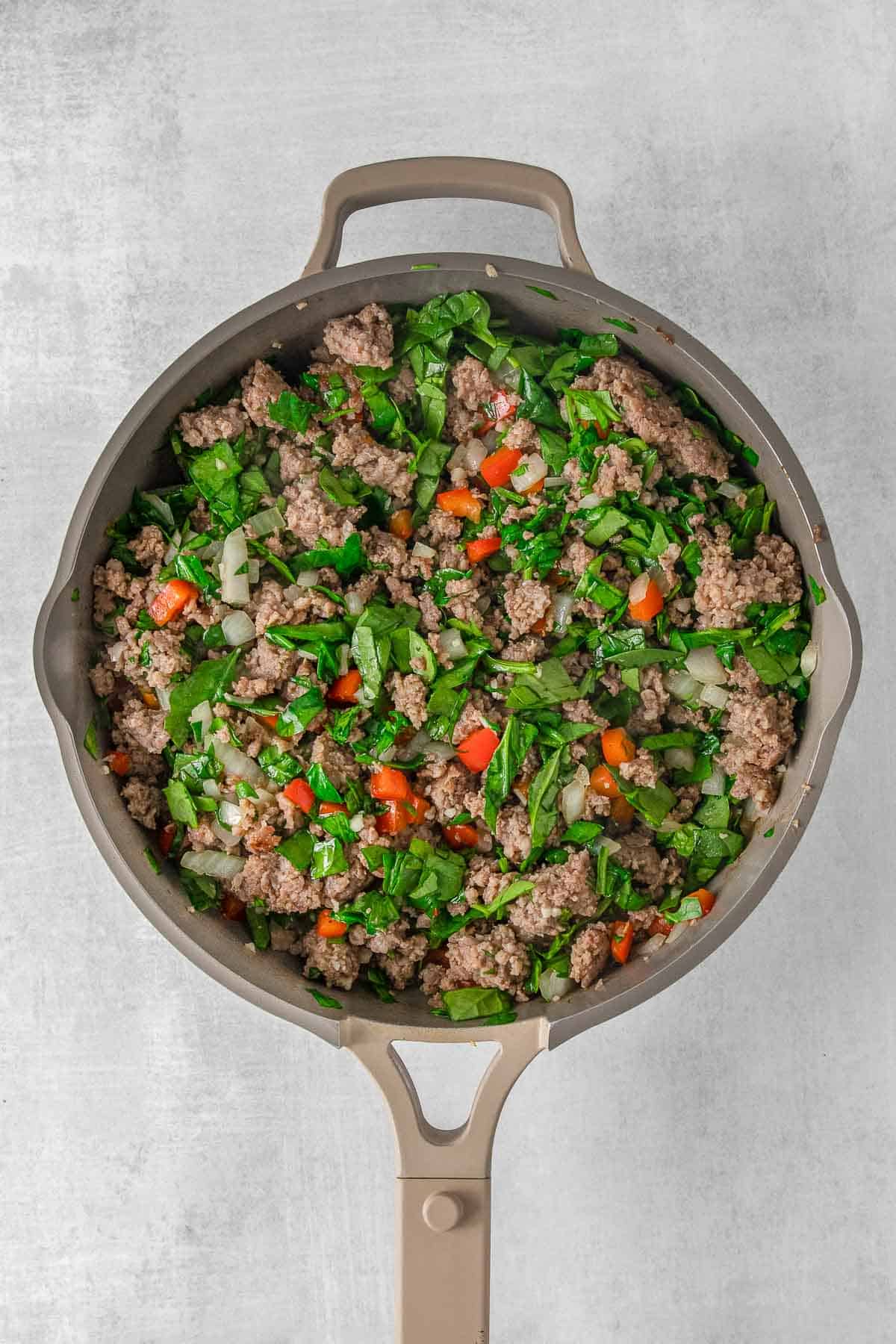 Large skillet with cooked sausage, red bell pepper, onion and garlic mixture topped with spinach.