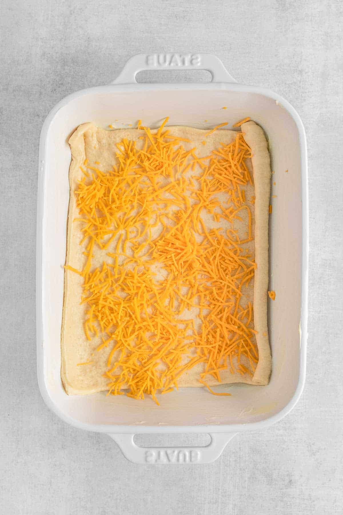 White casserole dish with unbaked crescent roll covered in shredded cheese.