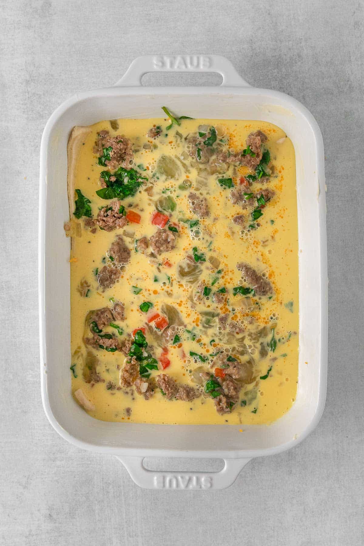White casserole dish with unbaked egg casserole mixture with sausage, spinach, red pepper and cheese.