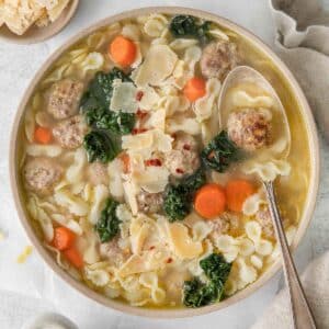 closeup of white serving bowl full homemade wedding soup with meatballs, kale, carrots and pasta with a spoon on the side of the bowl.