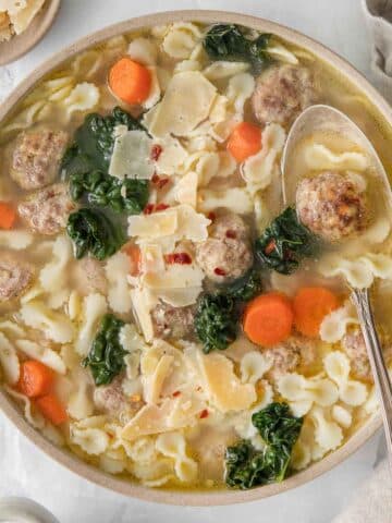 closeup of white serving bowl full homemade wedding soup with meatballs, kale, carrots and pasta with a spoon on the side of the bowl.