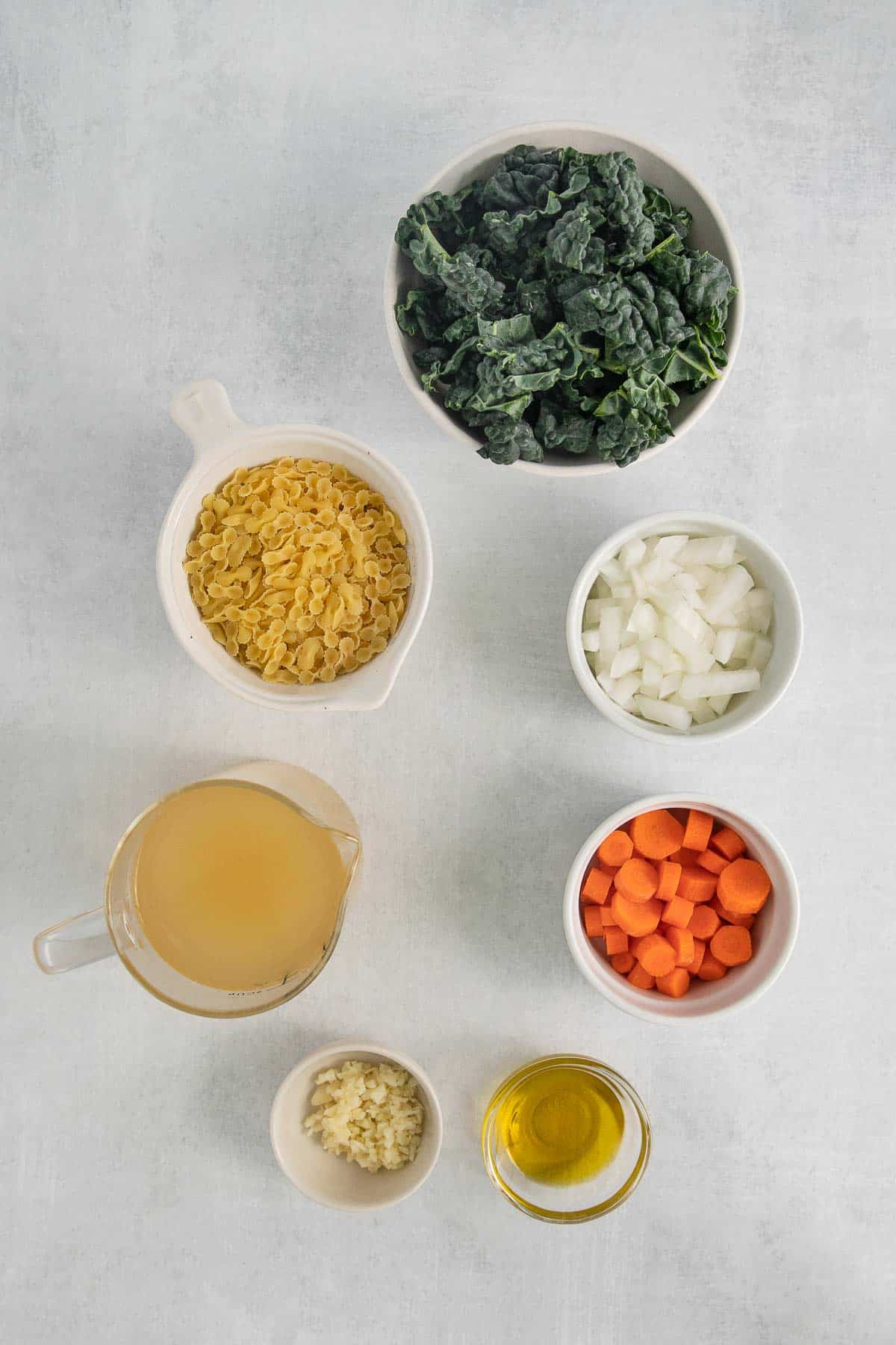 several bowls with ingredients for Italian wedding soup - kale, sliced carrots, diced onions, broth, minced garlic, oil, and pasta.