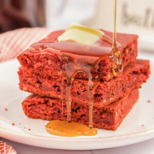 Stack of three red velvet pancakes with butter and maple syrup drizzle on a white plate.