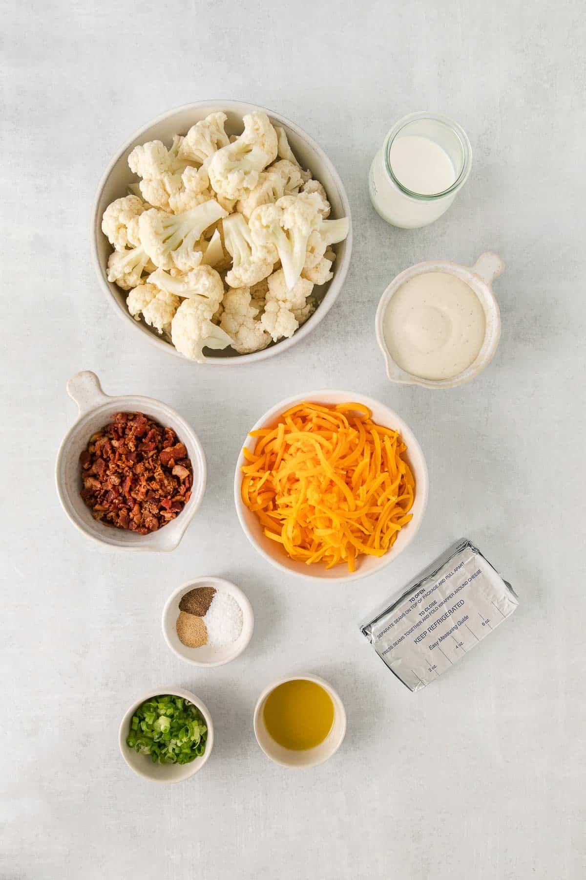 Several bowls with ingredients for Cheesy Cauliflower Casserole - Cauliflower, Cheese, Cream Cheese, Olive Oil, Seasoning Mix, Ranch, Milk, Bacon, Green Onion.