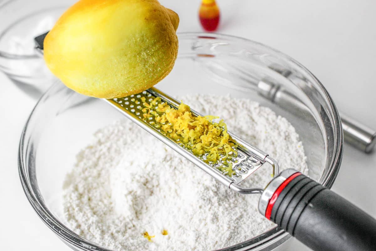 Lemon being zested over glass bowl of flour mixture.