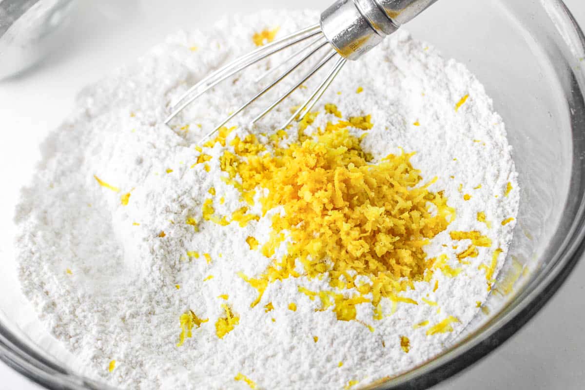 Glass mixing bowl of flour mixture and lemon zest with whisk in bowl.